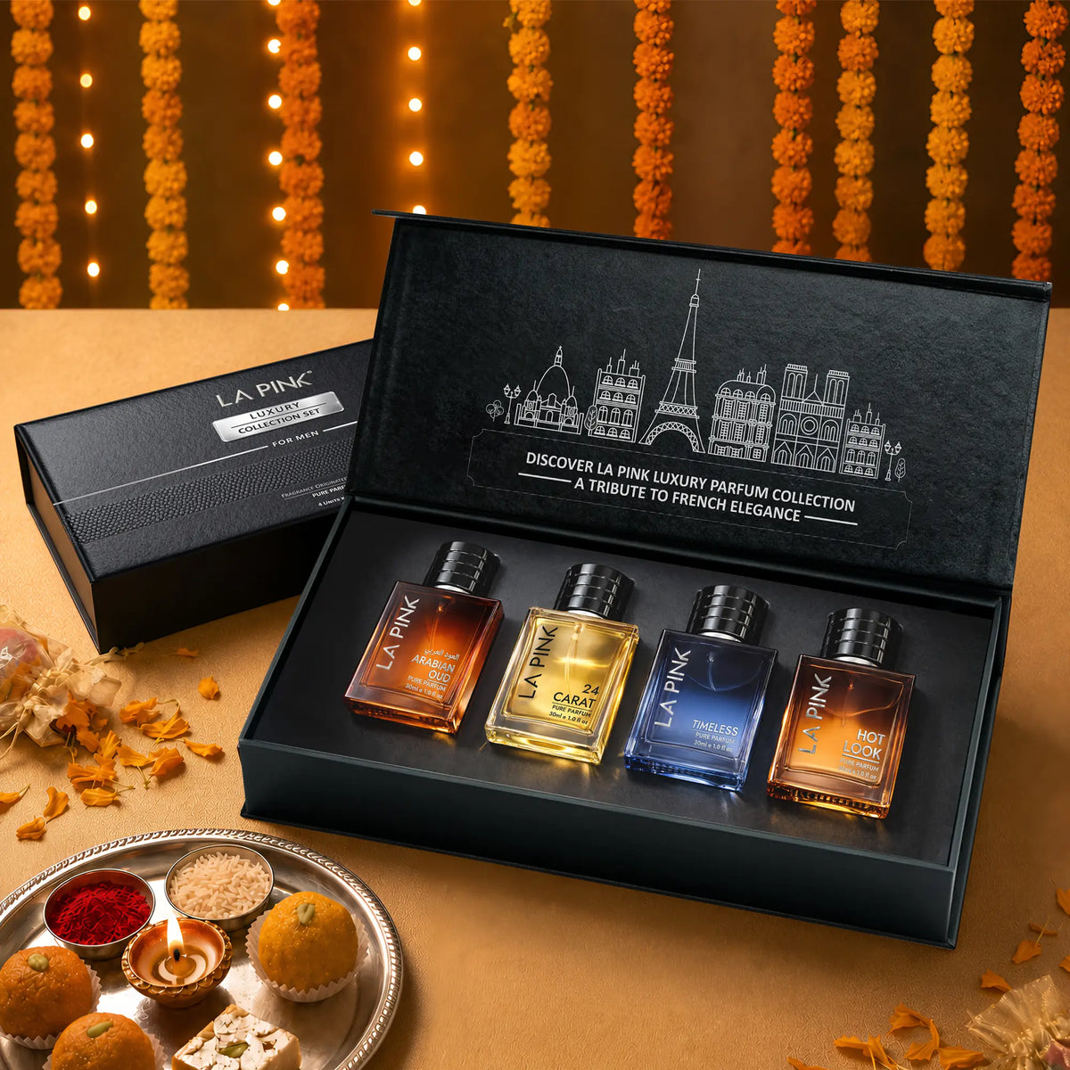LA PINK Luxury Parfum Collection gift set in an open black presentation box, featuring four miniature glass perfume bottles with amber, gold, blue, and orange liquids. The set is styled on a warm beige surface with decorative marigold garlands and sweets in the background.