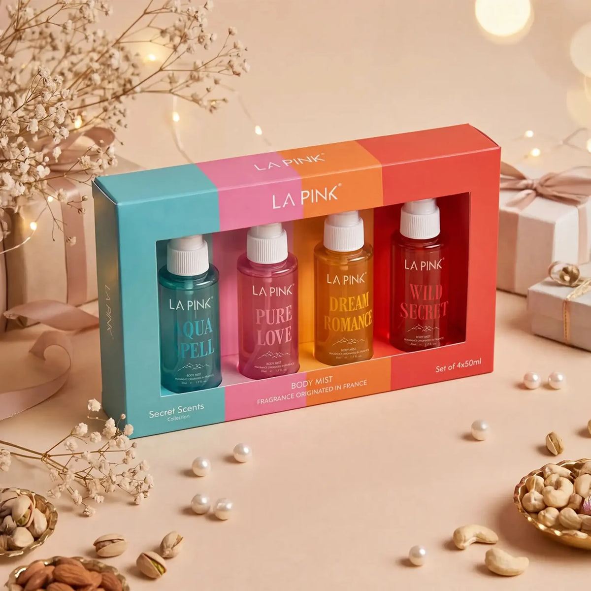 LA PINK body mist set in a colorful box with four different scents: Aqua Pell, Pure Love, Dream Romance, and Wild Secret. The set is styled on a beige surface with decorative dried flowers, pearls, and gift boxes in the background.