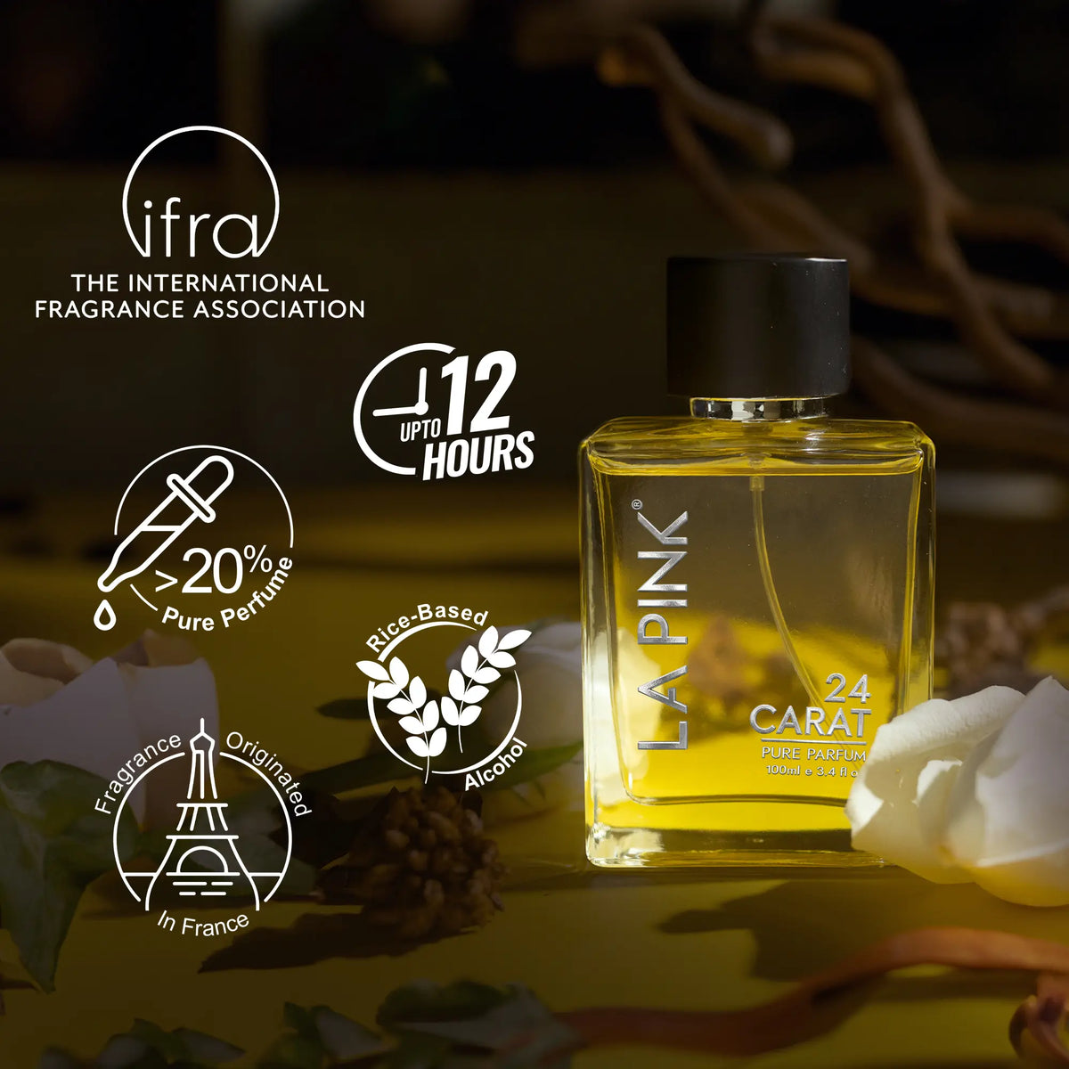 A bottle of LA PINK 24 CARAT perfume displayed on a dark, atmospheric background with white rose petals and decorative text badges from the IFRA (International Fragrance Association). The product is a rectangular glass bottle filled with golden-yellow liquid, topped with a black cap, and features white text.

