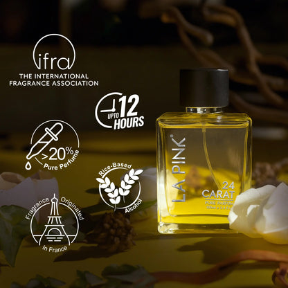A bottle of LA PINK 24 CARAT perfume displayed on a dark, atmospheric background with white rose petals and decorative text badges from the IFRA (International Fragrance Association). The product is a rectangular glass bottle filled with golden-yellow liquid, topped with a black cap, and features white text.

