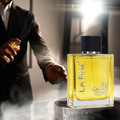 A bottle of LA PINK 24 CARAT Full Perfume Oil with golden-yellow liquid and a black cap is shown in the foreground, with a man in a dark suit holding and spraying a matching perfume bottle in a smoky, dramatic setting.