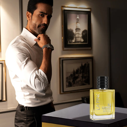 A man in a white dress shirt stands in a dimly lit room next to a bottle of La Pink by Gholap &amp; Co fragrance displayed on a table. The bottle is clear with yellow liquid and a black cap.

