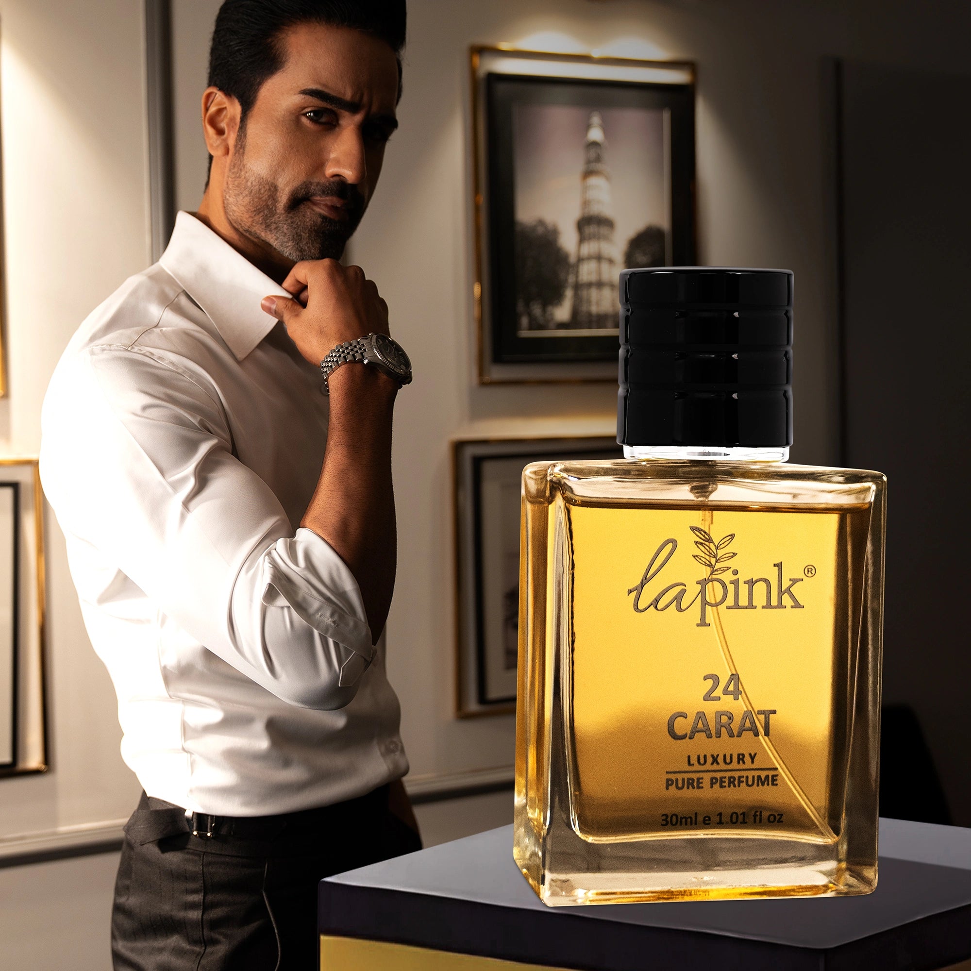 Man in a white shirt with a lapink 24 Carat perfume bottle in a room with framed pictures on the wall.