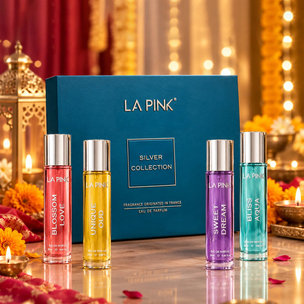 LA PINK Silver Collection fragrance gift set featuring four 10ml travel spray bottles in pink, yellow, purple, and blue-green, displayed in front of a blue gift box on a reflective surface with warm bokeh lights and candles.

