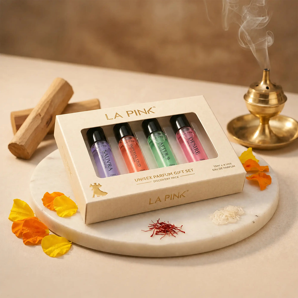 LA PINK Unisex Parfum Gift Set containing four 10ml travel atomizers in purple, orange, green, and pink scents, presented in a cream-colored box on a marble tray with decorative petals and incense.

