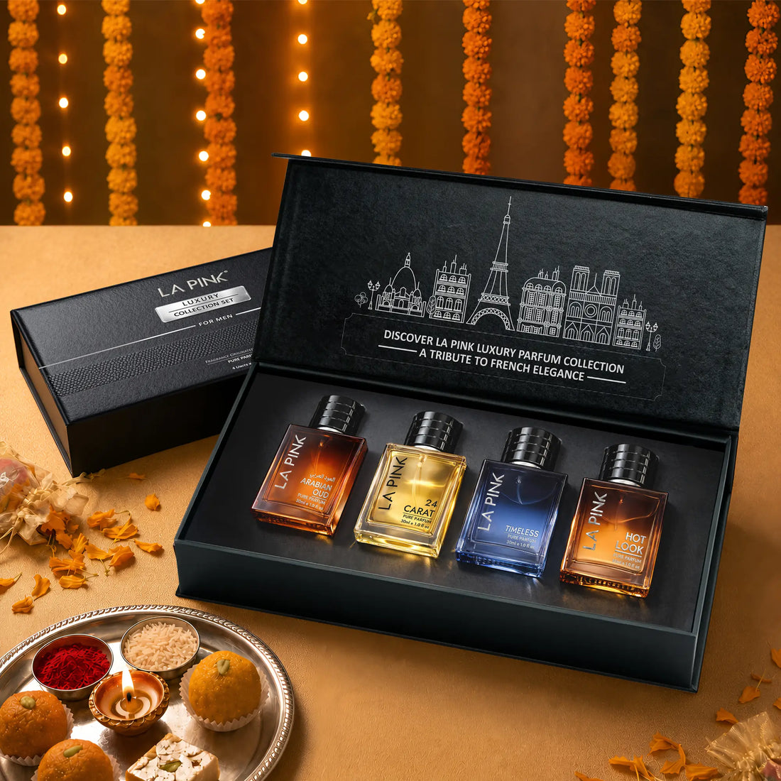LA PINK Luxury Parfum Collection gift set in an open black presentation box, featuring four miniature glass perfume bottles with amber, gold, blue, and orange liquids. The set is styled on a warm beige surface with decorative marigold garlands and sweets in the background.

