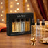 LA PINK pure perfume set with a black 4-pack box and four individual spray bottles displayed on a wooden table with warm bokeh lights in the background.

