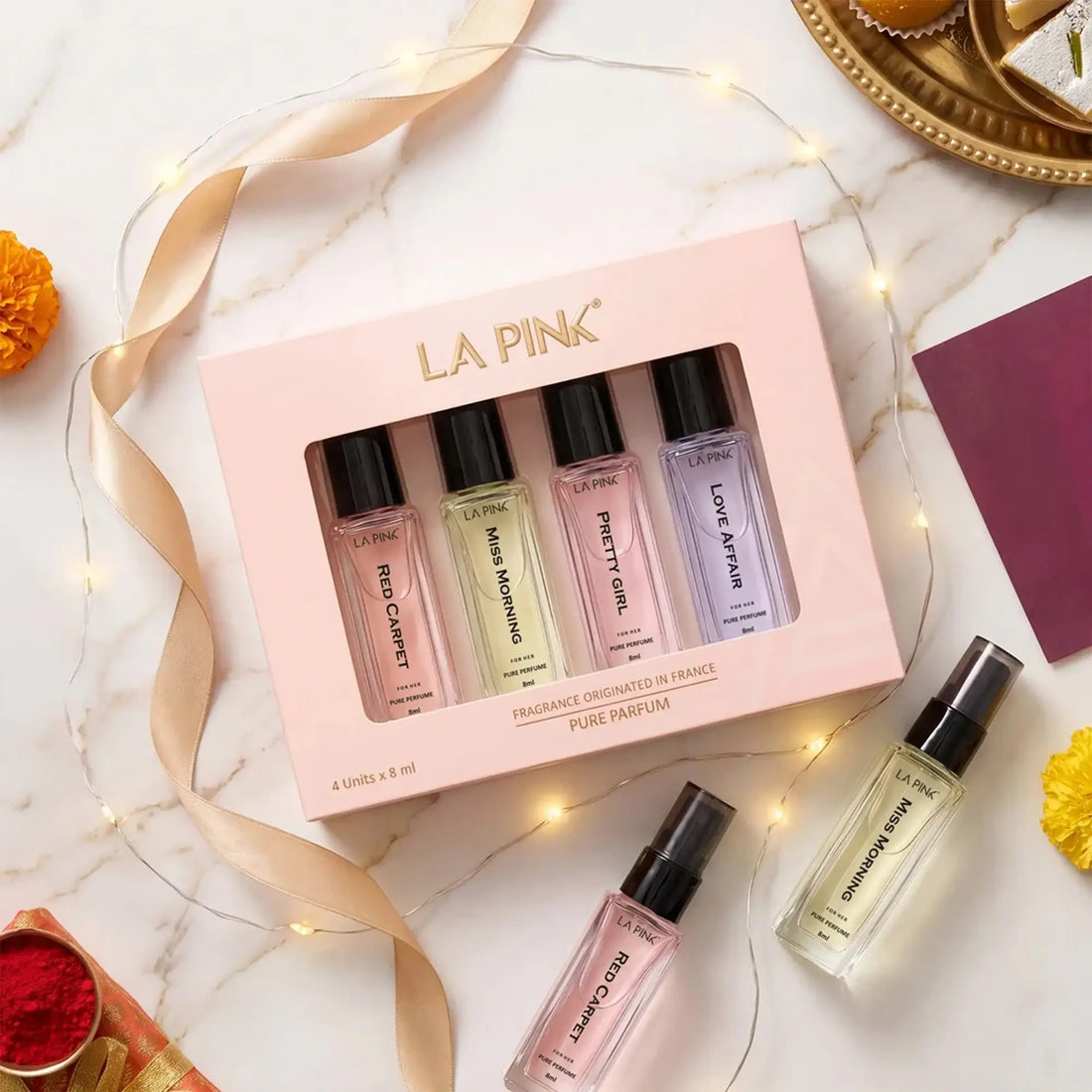 A boxed fragrance gift set from LA PINK on a white marble surface with fairy lights and ribbon. The pale pink box contains four small perfume bottles, and two additional bottles are placed outside the box.

